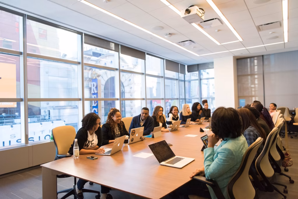 Group of colleagues in a modern conference room seated around a long table with laptops, engaged in a business meeting near floor-to-ceiling windows.